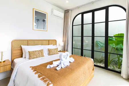Spacious tropical bedroom with rattan headboard, king bed adorned with white linens and towel swan, arched black-framed windows overlooking lush greenery.